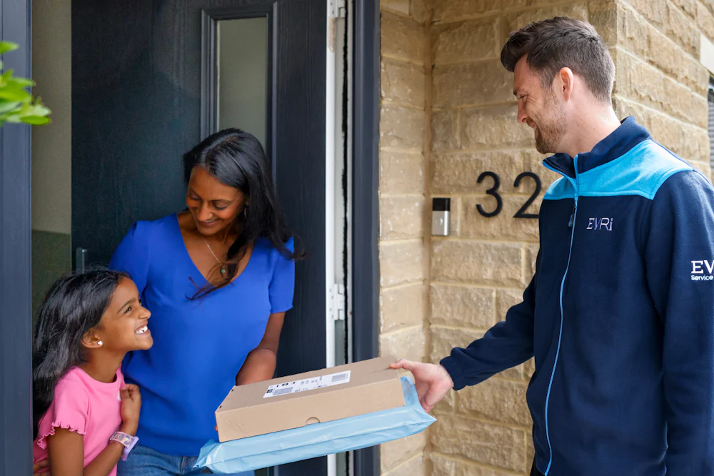 An Evri Courier handing two parcels to a mother and child on their doorstep as they look to eachother excited. Another child is playing with a ball and bat in the background.