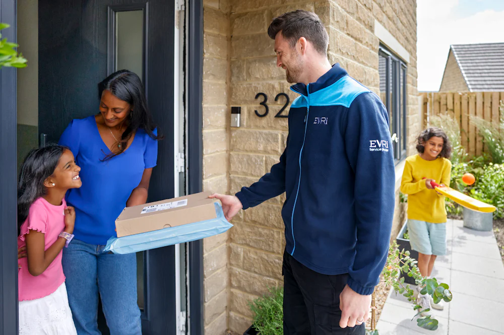 An Evri Courier handing two parcels to a mother and child on their doorstep as they look to eachother excited. Another child is playing with a ball and bat in the background.