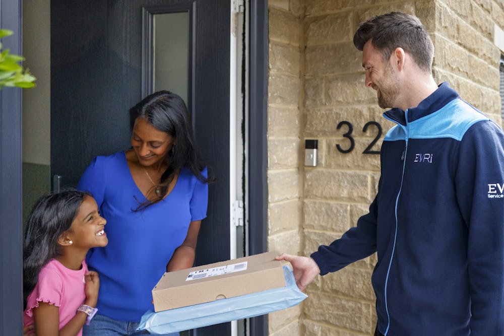 An Evri Courier handing two parcels to a mother and child on their doorstep as they look to eachother excited. Another child is playing with a ball and bat in the background.