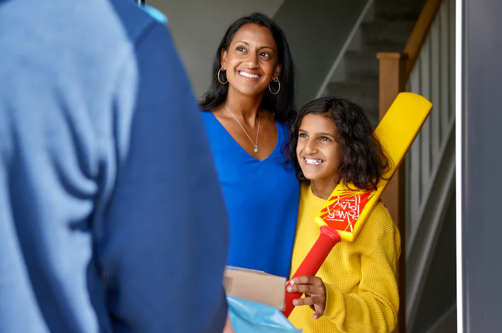An Evri Courier is handing two parcels to a mother and child as they look very pleased towards the courier. The child is holding a cricket bat.