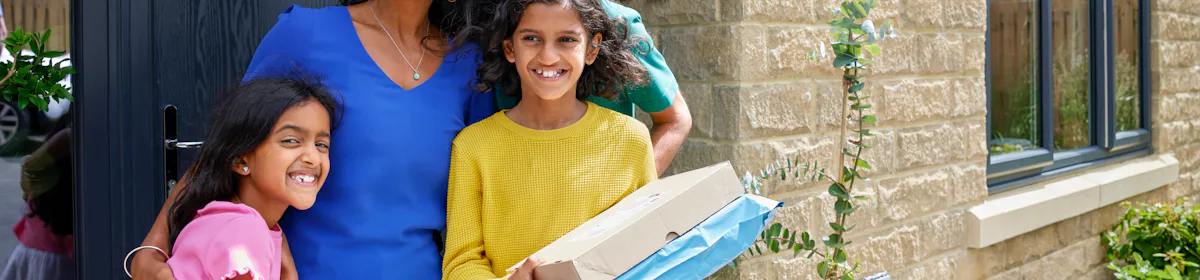 A family of four pose smiling in front of their front door holding two parcels. The family consists of a father, mother and two daughters.