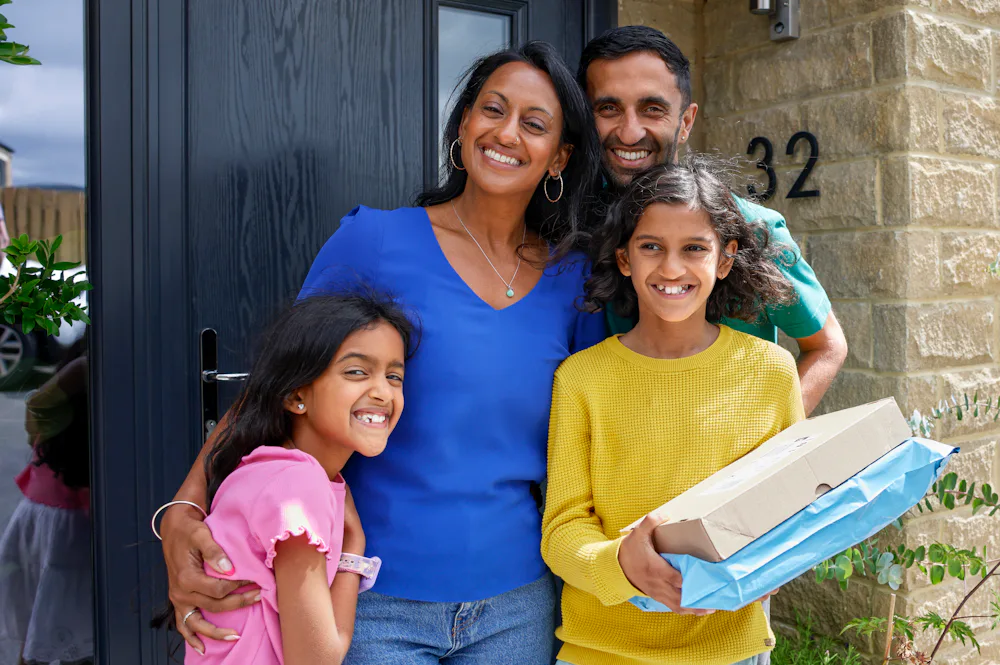 A family of four pose smiling in front of their front door holding two parcels. The family consists of a father, mother and two daughters.
