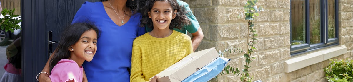 A family of four pose smiling in front of their front door holding two parcels. The family consists of a father, mother and two daughters.