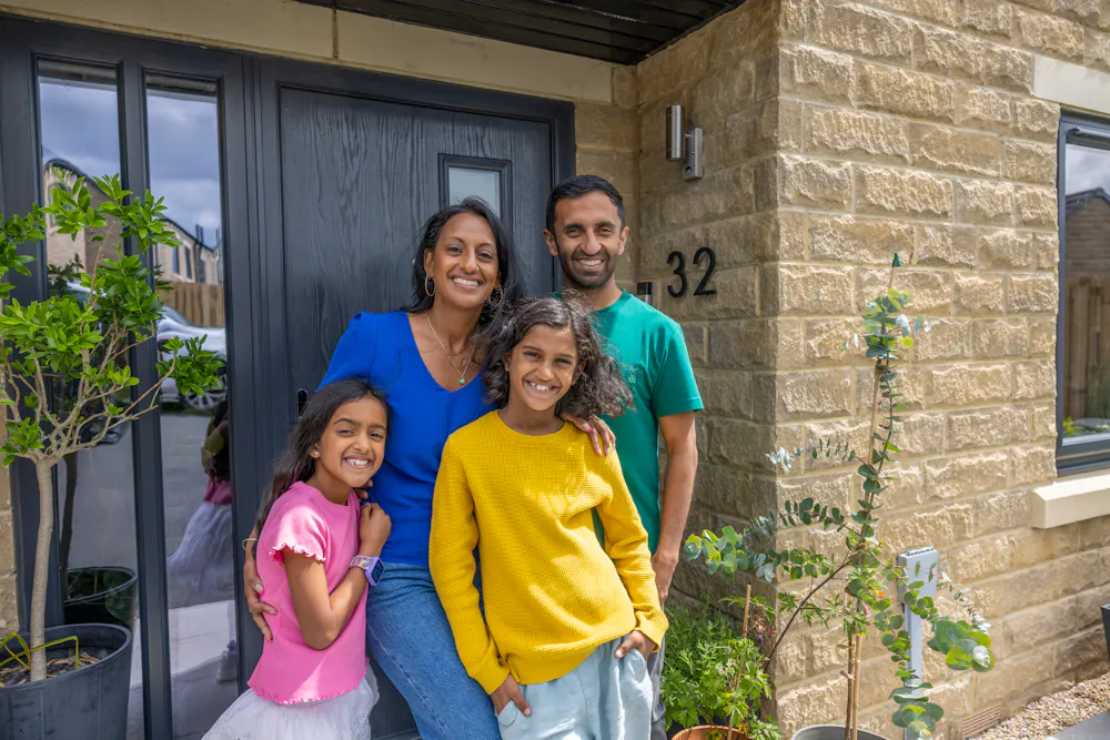 A family of four pose smiling in front of their front door. The family consists of a father, mother and two daughters.