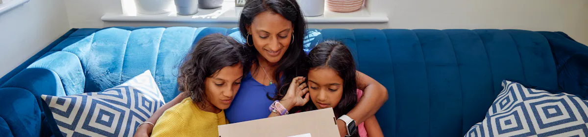 A mother and her two children open up a parcel. They are sat cuddled up on a sofa in their living room.
