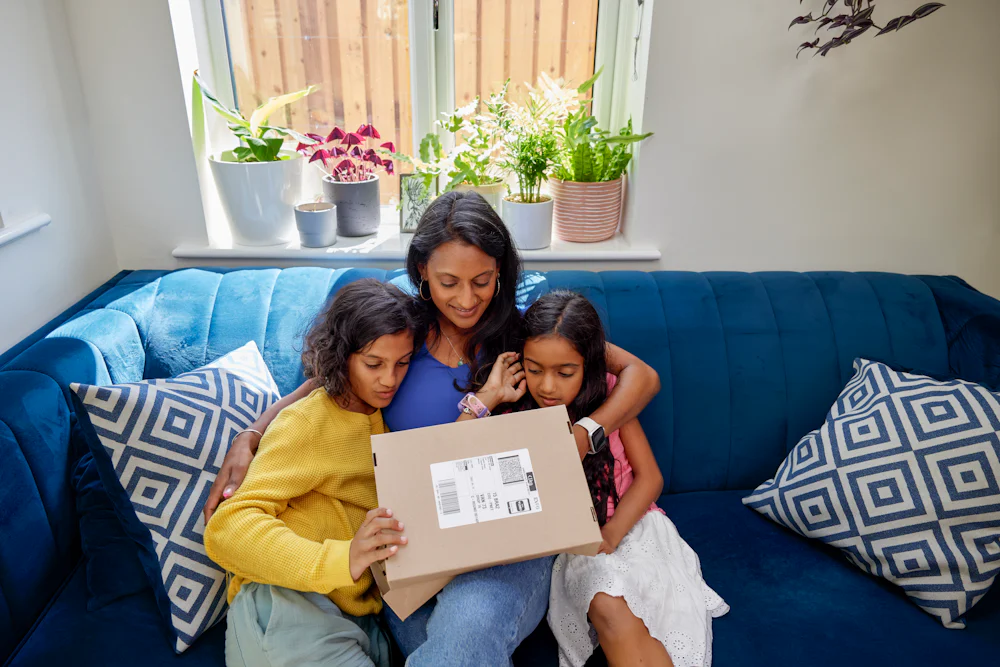 A mother and her two children open up a parcel. They are sat cuddled up on a sofa in their living room.