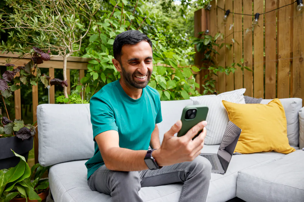 A man is sat outdoors on a sofa bench recording an Evri video on his mobile device.