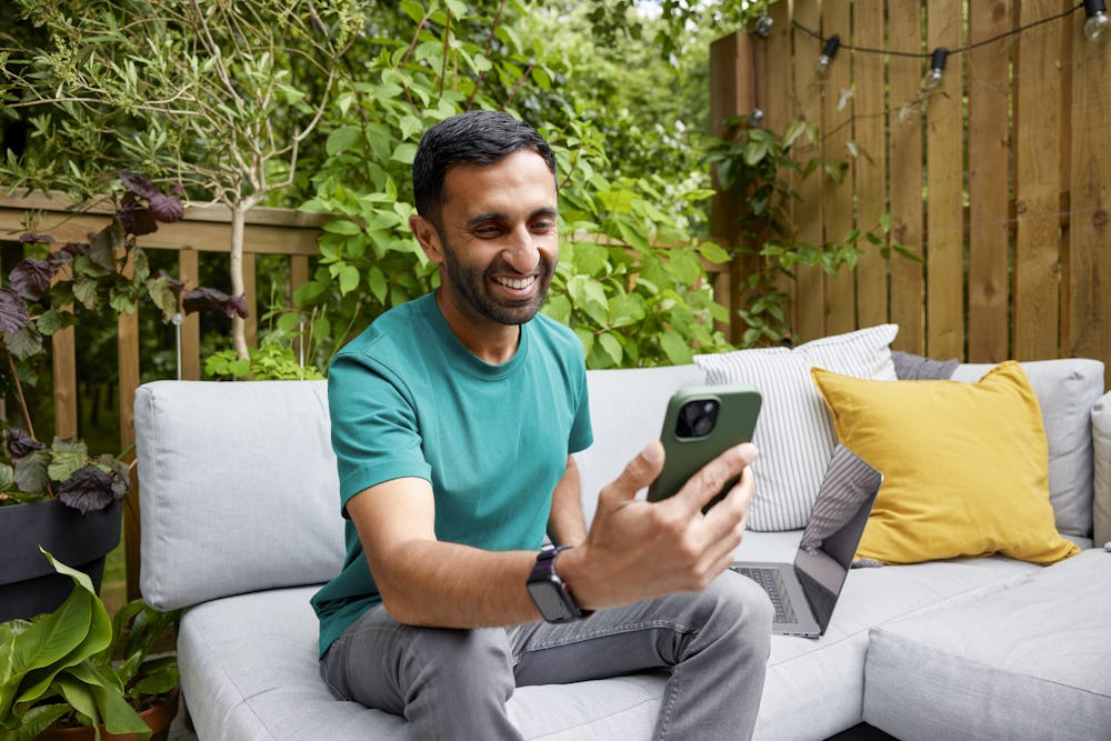 A man is sat outdoors on a sofa bench recording an Evri video on his mobile device.