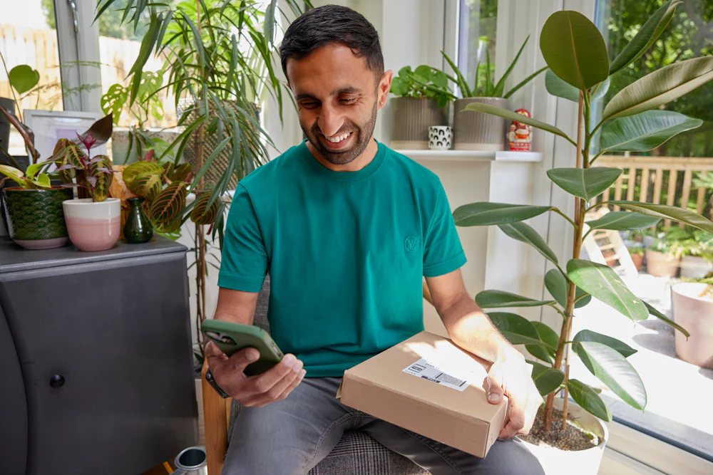 A man is sat with a parcel on his lap, watching the accompanying Evri video on his mobile phone. He is laughing as he watches the video.