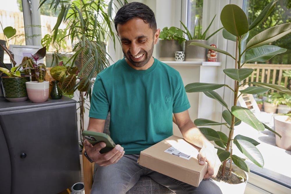 A man is sat with a parcel on his lap, watching the accompanying Evri video on his mobile phone. He is laughing as he watches the video.
