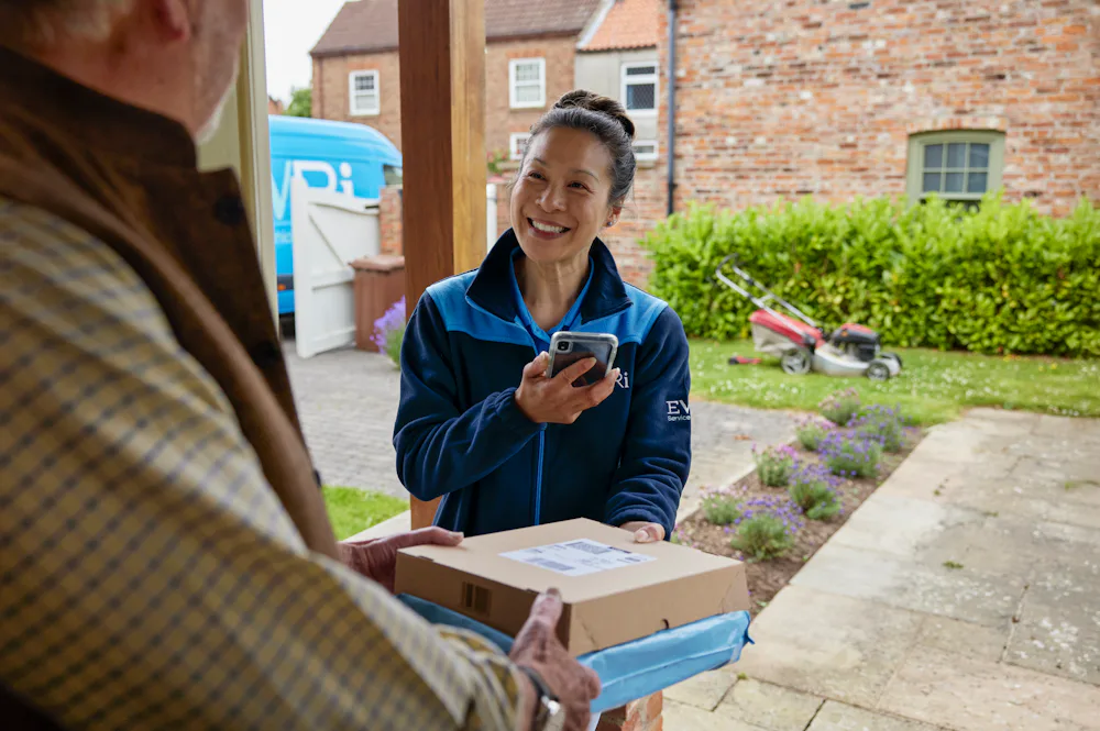 A handful of parcels are being handed to a recipient at the door. A blue Evri van can be seen in the background.