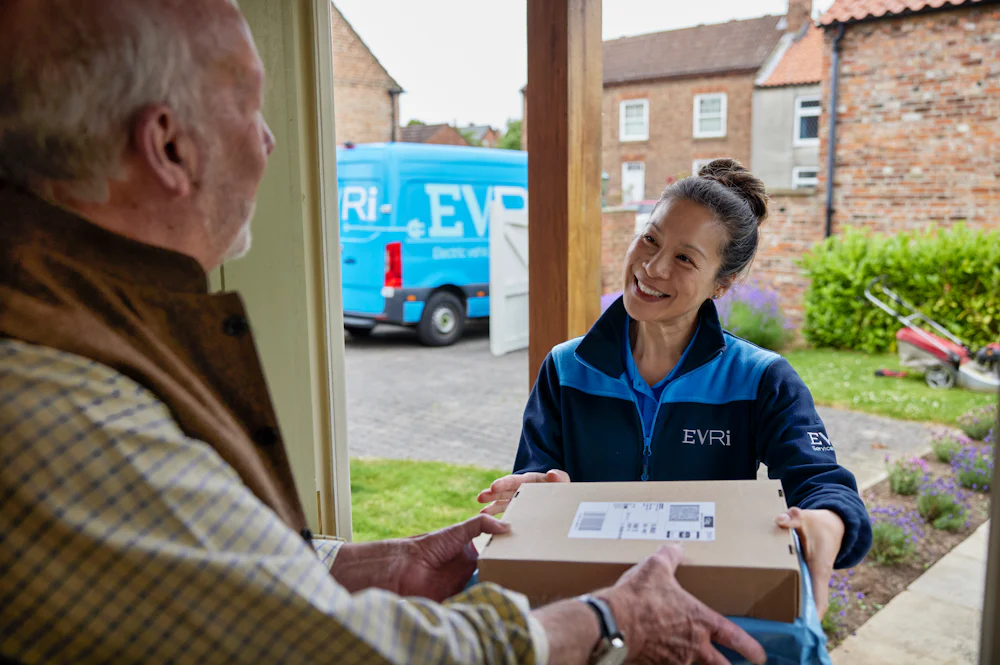 A handful of parcels are being handed to a recipient at the door. A blue Evri van can be seen in the background.