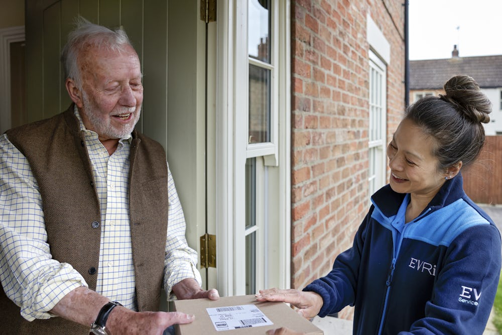 A handful of parcels are being handed to a recipient at the door. Both the courier and recipient are clearly visible