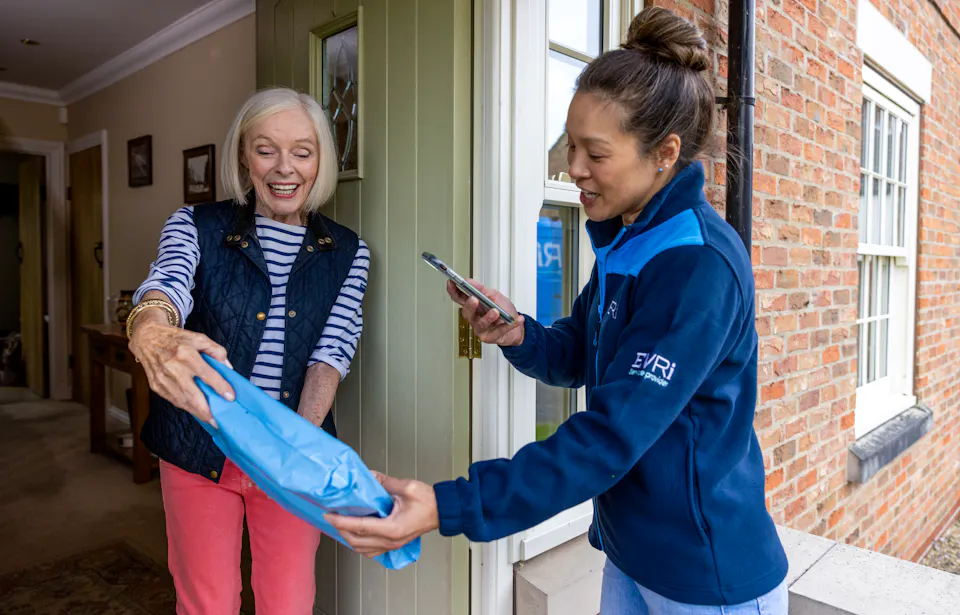 A parcel in waterproof blue packaging is being photographed by a courier as proof of delivery. Both the courier and recipient are clearly visible