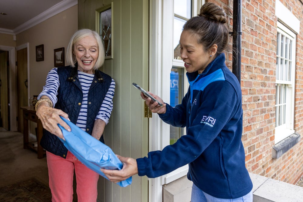A parcel in waterproof blue packaging is being photographed by a courier as proof of delivery. Both the courier and recipient are clearly visible