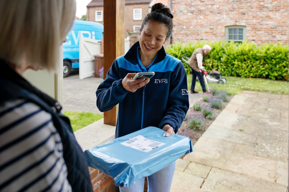 A parcel in waterproof blue packaging is being photographed by a courier as proof of delivery. A blue Evri van can be seen in the background.