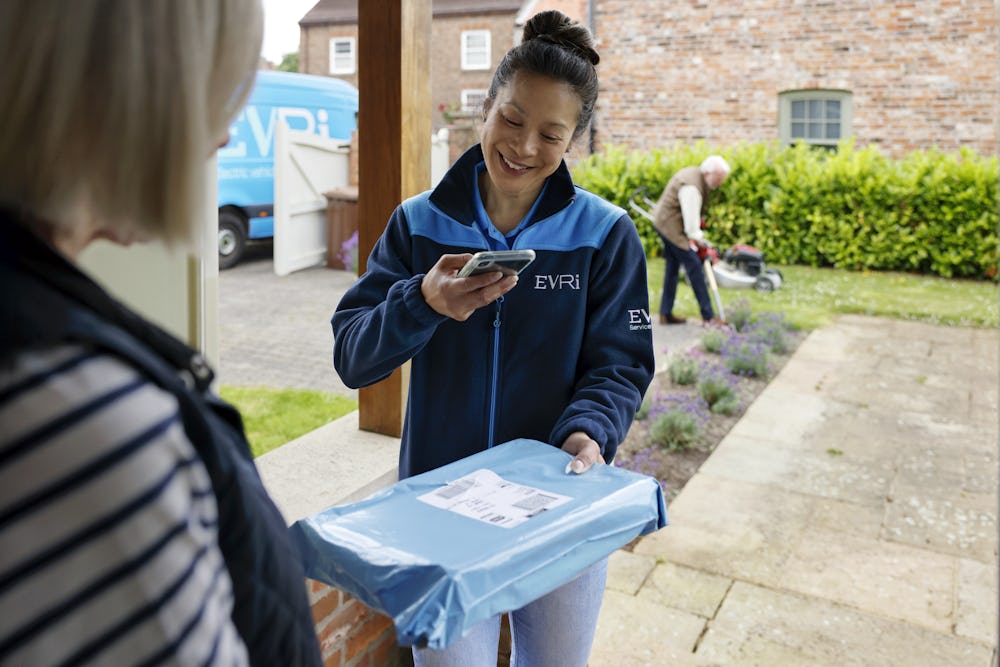 A parcel in waterproof blue packaging is being photographed by a courier as proof of delivery. A blue Evri van can be seen in the background.