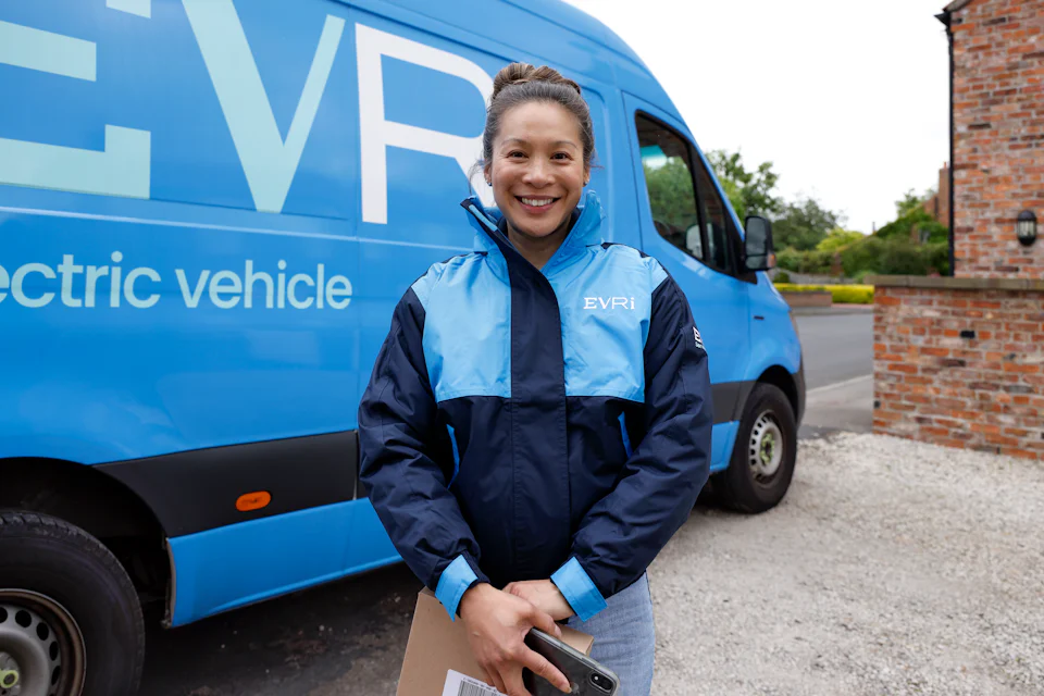 A female courier dressed in an Evri uniform stands in front of a blue Evri van. She is posing with a parcel in her hands.