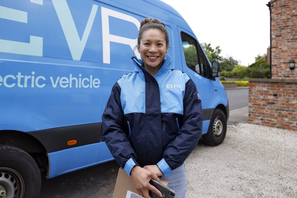 A female courier dressed in an Evri uniform stands in front of a blue Evri van. She is posing with a parcel in her hands.