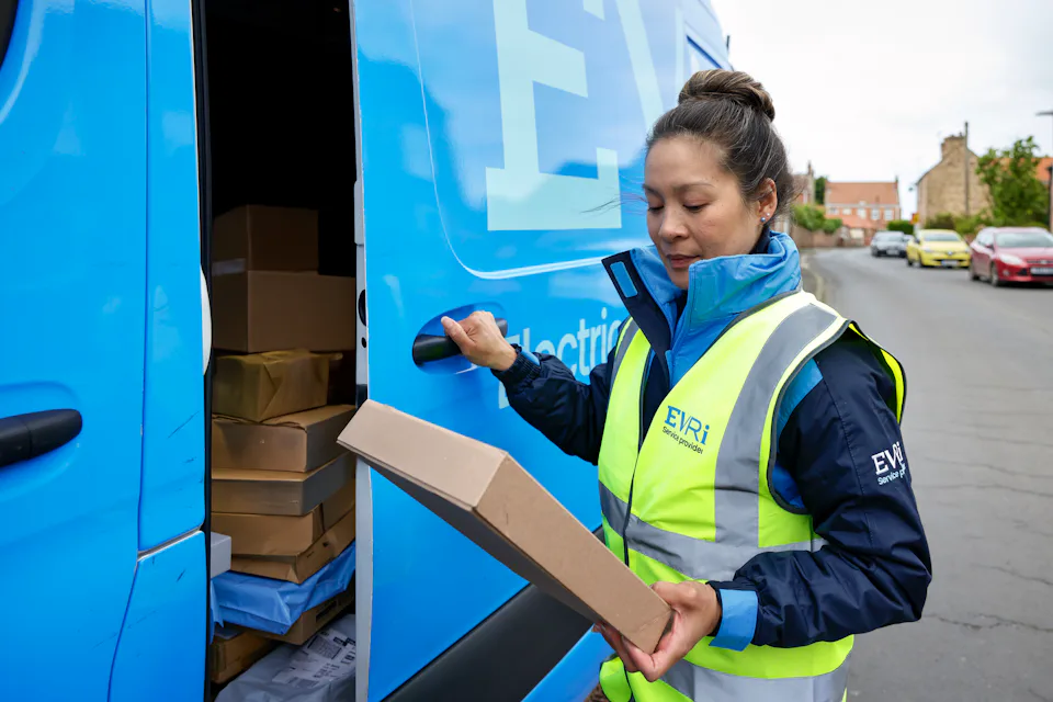 A female van driver dressed in an Evri banded hi-vis vest is loading a blue Evri van with parcels.