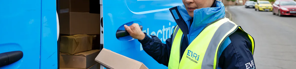 A female van driver dressed in an Evri banded hi-vis vest is loading a blue Evri van with parcels.