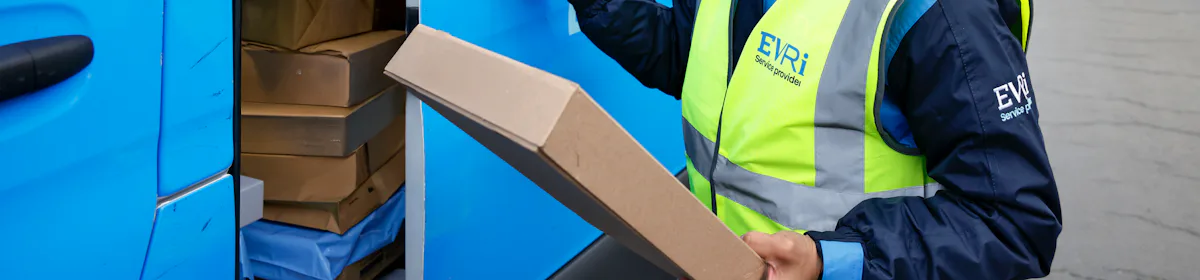 A female van driver dressed in an Evri banded hi-vis vest is loading a blue Evri van with parcels.