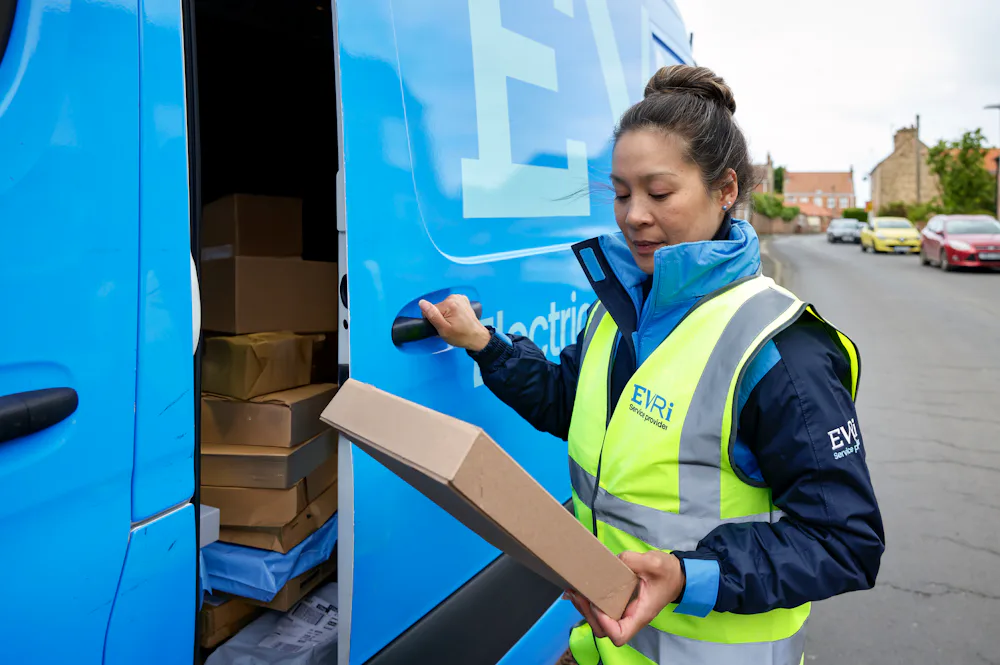 A female van driver dressed in an Evri banded hi-vis vest is loading a blue Evri van with parcels.