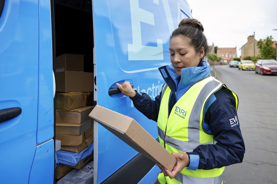 A female van driver dressed in an Evri banded hi-vis vest is loading a blue Evri van with parcels.