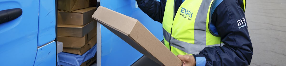 A female van driver dressed in an Evri banded hi-vis vest is loading a blue Evri van with parcels.