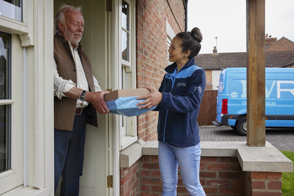 A handful of parcels are being handed to a recipient at the door. Both the courier and recipient are clearly visible