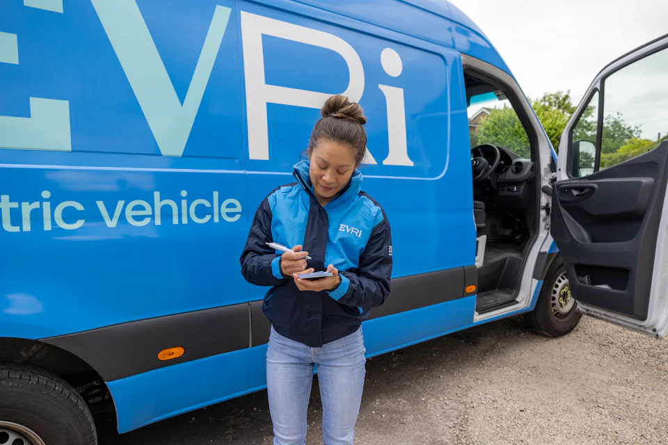 A female courier dressed in an Evri uniform stands in front of a blue Evri van with a handful of delivery slips.