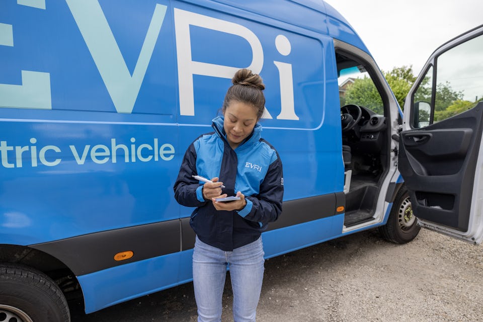 A female courier dressed in an Evri uniform stands in front of a blue Evri van with a handful of delivery slips.