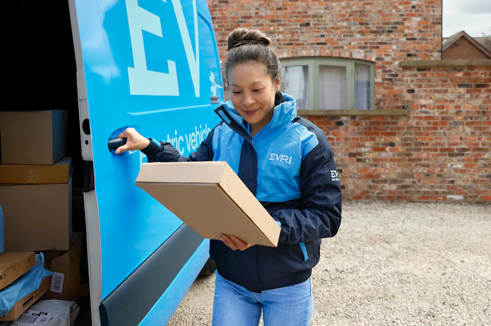 A female van driver dressed in an Evri banded vest is loading a blue Evri van with parcels.