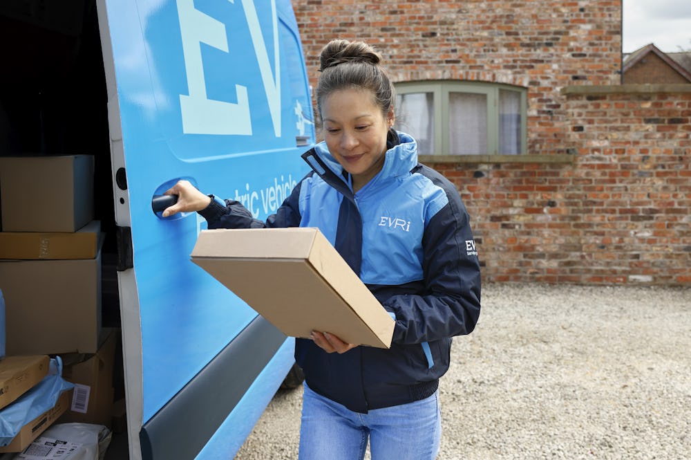 A female van driver dressed in an Evri banded vest is loading a blue Evri van with parcels.