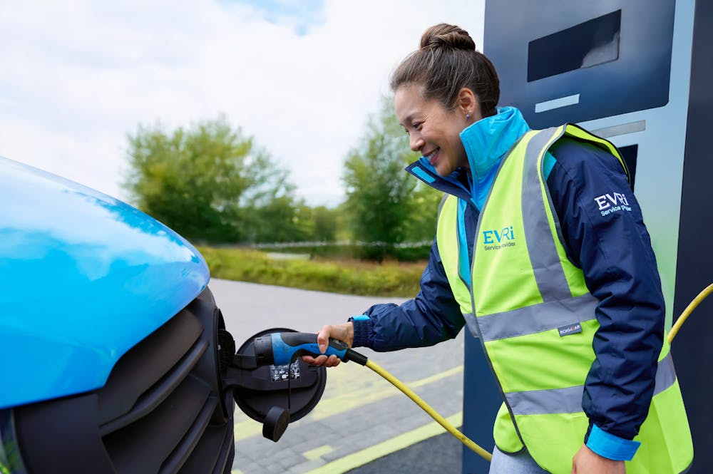 A female van driver dressed in an Evri branded hi-vis vest is charging a blue Evri electric van.