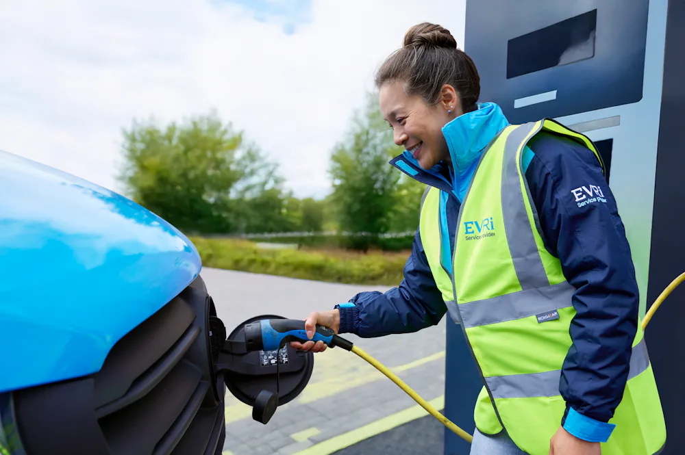 A female van driver dressed in an Evri branded hi-vis vest is charging a blue Evri electric van.