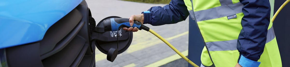 A female van driver dressed in an Evri branded hi-vis vest is charging a blue Evri electric van.
