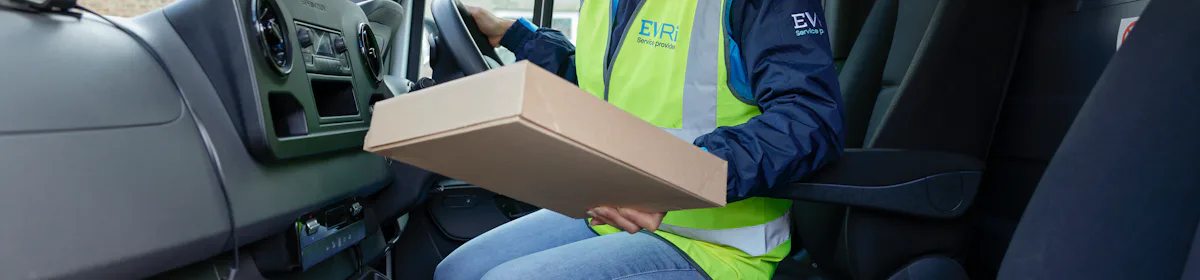 A female van driver dressed in an Evri branded hi-vis vest is sat in the cabin of her van. A pile of parcels is placed on the seat beside her.