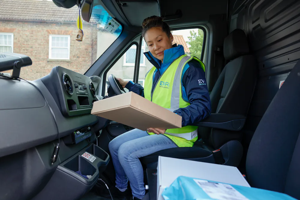 A female van driver dressed in an Evri branded hi-vis vest is sat in the cabin of her van. A pile of parcels is placed on the seat beside her.