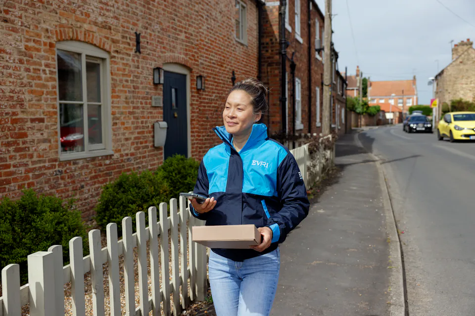 A female courier is walking up a quiet British street. A white picket fence can be seen beside her and she is holding a brown paper parcel in her hand.
