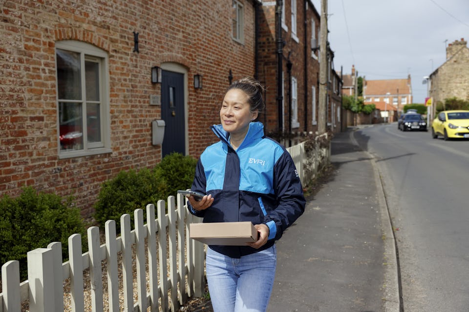 A female courier is walking up a quiet British street. A white picket fence can be seen beside her and she is holding a brown paper parcel in her hand.