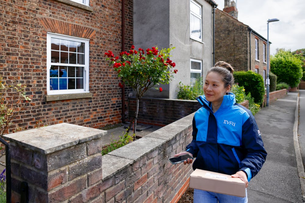 A female courier is walking up a quiet British street. A brick wall can be seen beside her and she is holding a brown paper parcel in her hand.