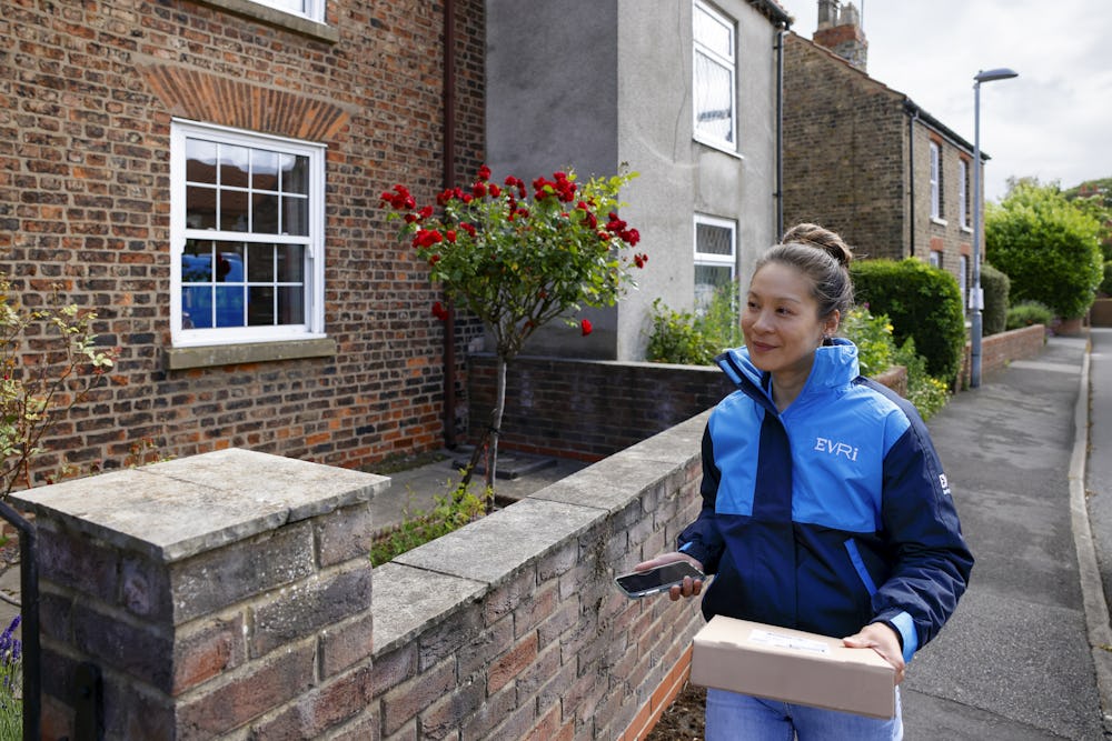 A female courier is walking up a quiet British street. A brick wall can be seen beside her and she is holding a brown paper parcel in her hand.
