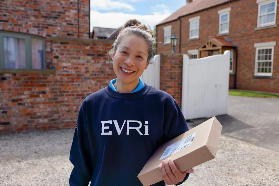 A female courier in an Evri branded sweater poses with a parcel on a driveway.