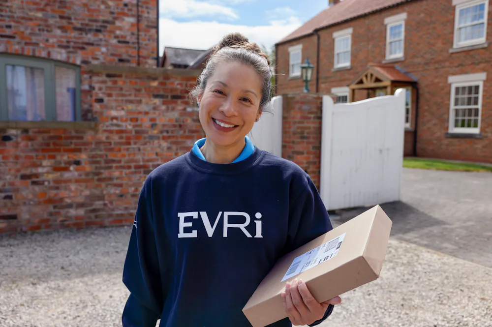 A female courier in an Evri branded sweater poses with a parcel on a driveway.