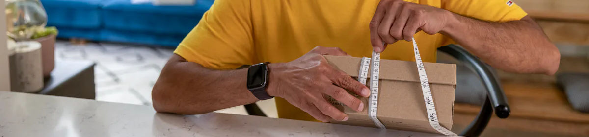 A father is measuring the circumference of a parcel box at his desk using measuring tape. His children are resting on the sofa behind him.