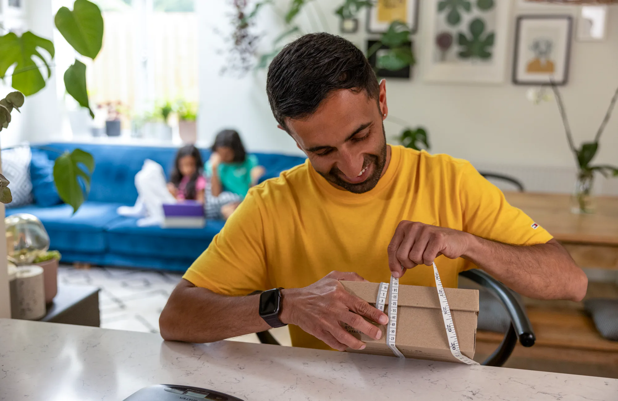 A father is measuring the circumference of a parcel box at his desk using measuring tape. His children are resting on the sofa behind him.