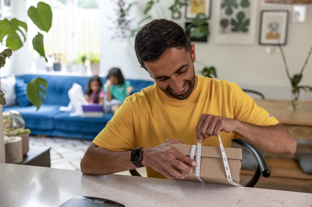 A father is measuring the circumference of a parcel box at his desk using measuring tape. His children are resting on the sofa behind him.