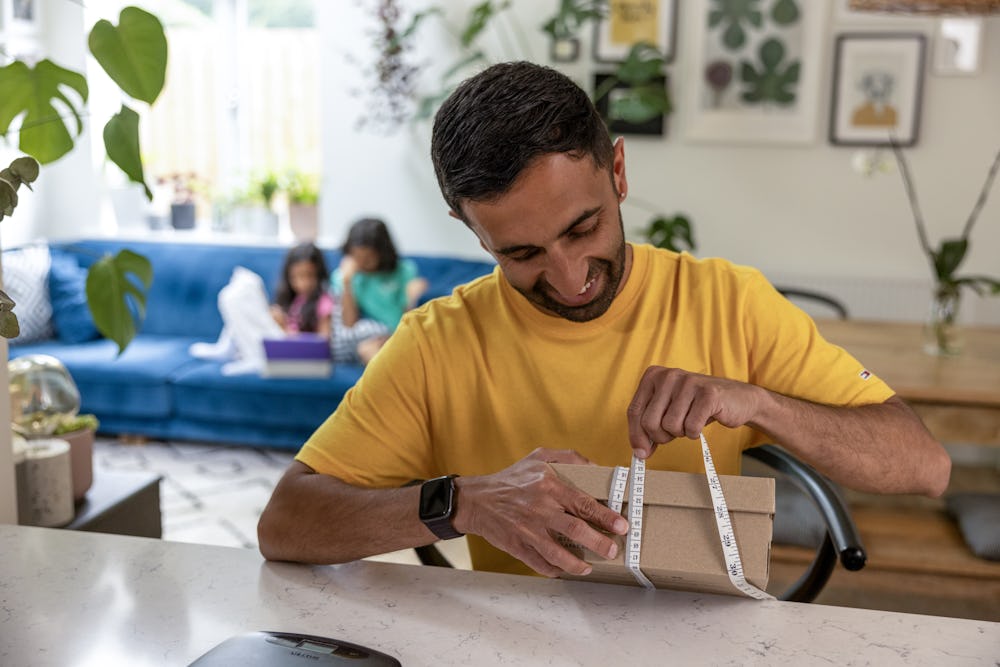 A father is measuring the circumference of a parcel box at his desk using measuring tape. His children are resting on the sofa behind him.