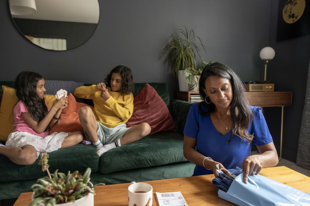 A mother is packaging a pair of trousers into a parcel bag with the delivery label ready to be attached. Her two children are sat playing cards on the sofa behind her
